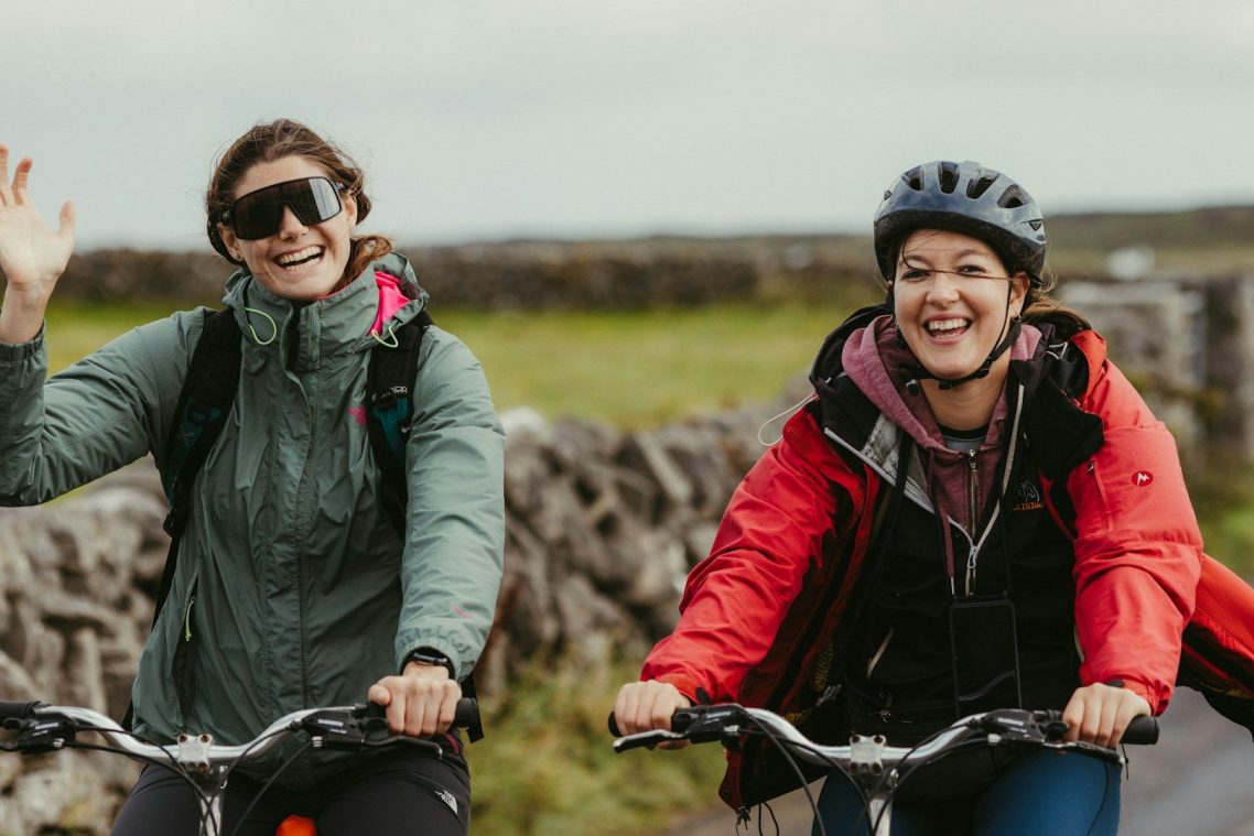 Comfy clothes, great ride. Two ladies riding their bikes on a road in front of a dry stone wall. One is waving, both are smiling, and wearing comfortable clothes.